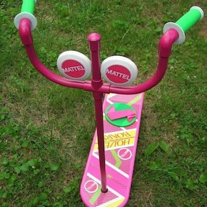 May include: A pink and green toy hoverboard with the words "Hover Board" and "Mattel" printed on it. The toy has two green handlebars and two white circular handles.