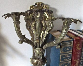 May include: Ornate gold-toned metal wall sconce with three candle holders. The sconce has a detailed, intricate design with a floral motif.