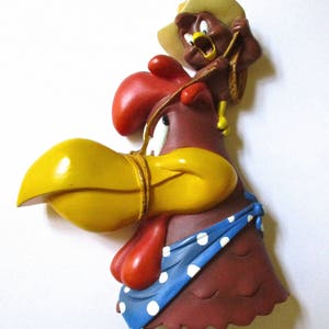 May include: A brown rooster figurine with a yellow beak and red wattle, wearing a blue bandana with white polka dots. A smaller brown rooster figurine with a yellow beak and a cowboy hat is perched on top of the larger rooster.