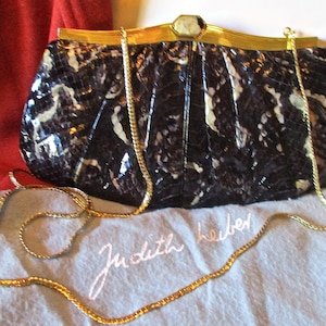 May include: A black and white snakeskin clutch purse with a gold chain strap and a gold clasp. The purse has a Judith Leiber label.