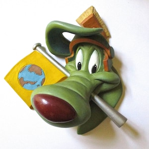 Puede incluir: Una figura de cabeza de perro verde de estilo de dibujos animados con una bandera amarilla que presenta un globo terráqueo azul y marrón. El perro lleva un casco marrón y naranja con un cepillo en la parte superior.