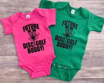 Può includere: Due body per neonati a maniche corte, uno rosa e uno verde. I body hanno un testo nero che recita "Future Disc Golf Buddy!" e un'immagine grafica nera di un cestino da disc golf.