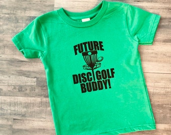 Può includere: Una maglietta verde brillante con la scritta nera "FUTURE DISC GOLF BUDDY!". La grafica presenta un canestro da disc golf e un disco. La maglietta è realizzata in un materiale morbido ed è probabilmente per un bambino.