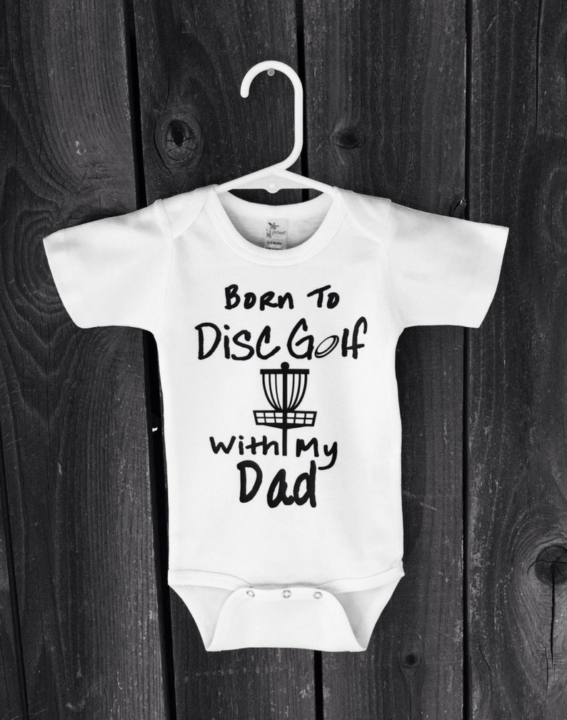 May include: White baby onesie with black text that reads "Born to Disc Golf with my Dad" and a black silhouette of a disc golf basket.