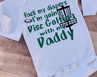 Può includere: Body bianco a maniche lunghe per neonato con testo verde che recita "Pack my diapers I'm going Disc Golfing with my Daddy". Accanto al testo è raffigurato un cestino da disc golf in bianco e nero.