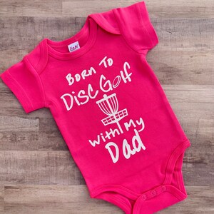 May include: Pink baby onesie with white text that reads "Born to Disc Golf with my Dad" and a white disc golf basket graphic.