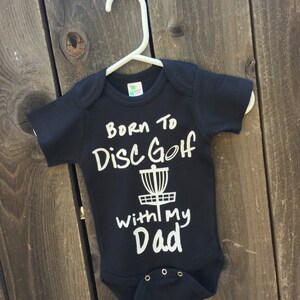 May include: Black baby onesie with white text that reads "Born to Disc Golf with my Dad" and a white disc golf basket graphic.