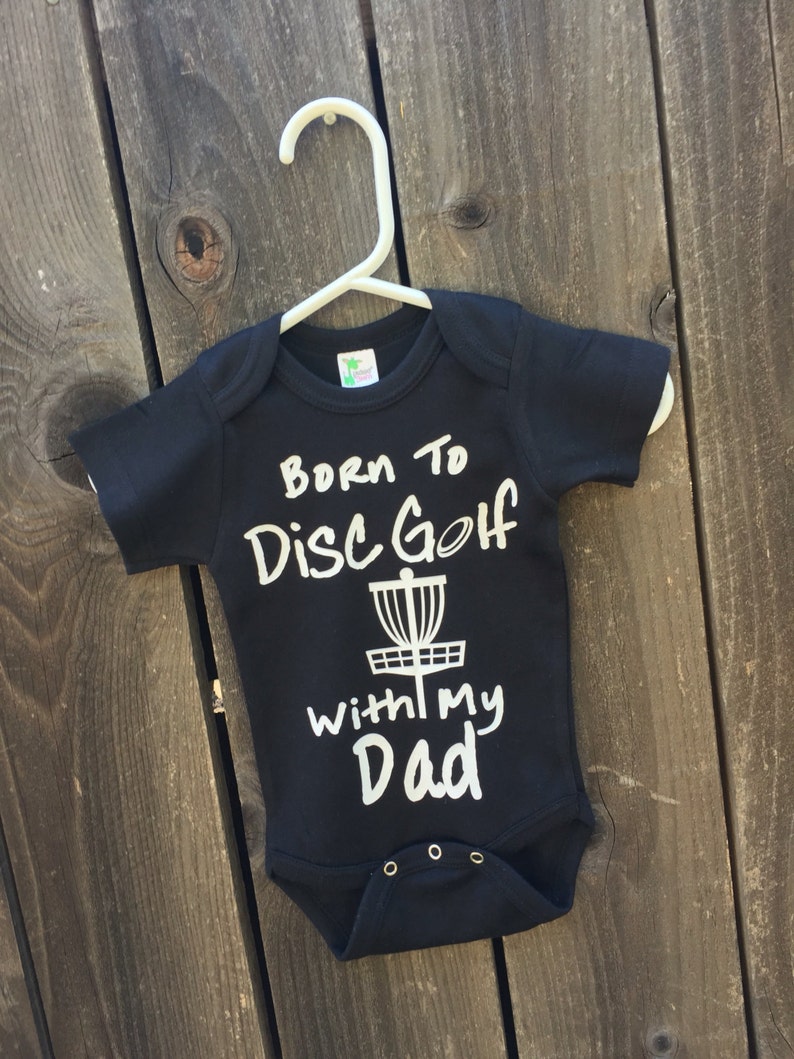 May include: Black baby onesie with white text that reads "Born to Disc Golf with my Dad" and a white disc golf basket graphic.