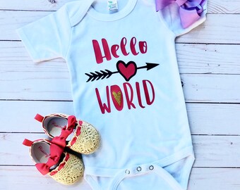 Può includere: Un body bianco per neonato con il testo "Hello World" in rosso con un cuore glitterato dorato e una freccia nera. Il body ha maniche corte e bottoni a pressione sul fondo. Nell'immagine sono presenti anche un fiocco viola e un paio di scarpine per neonato dorate e rosse.