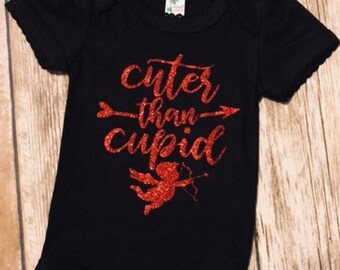 Può includere: Body nero per neonato con testo in glitter rosso che dice "cuter than cupid" e una silhouette di cupido in glitter rosso.