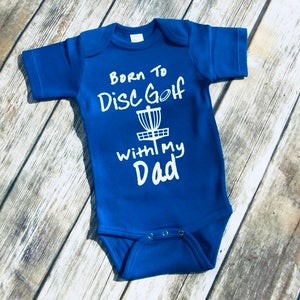 May include: Blue baby onesie with white text that reads "Born to Disc Golf with my Dad" and a white silhouette of a disc golf basket.