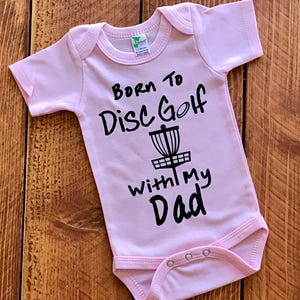 May include: Pink baby onesie with black text that reads "Born to Disc Golf with my Dad" and a black silhouette of a disc golf basket.