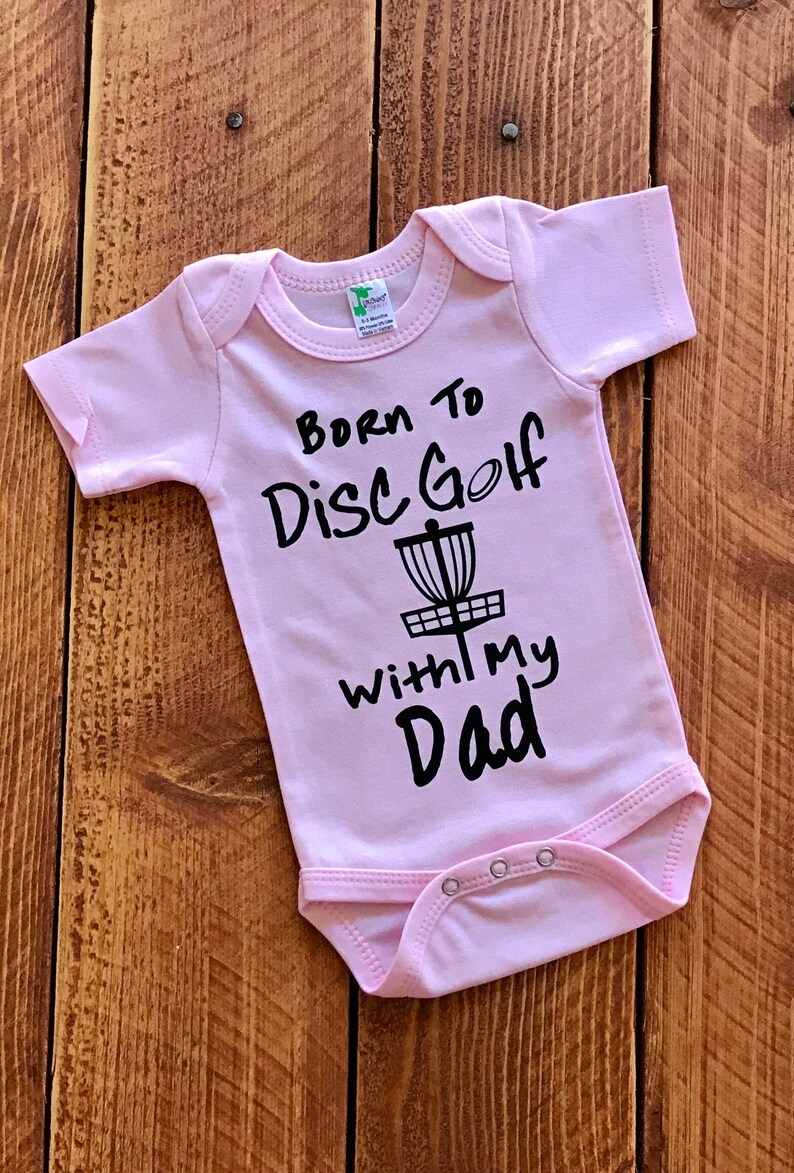 May include: Pink baby onesie with black text that reads "Born to Disc Golf with my Dad" and a black silhouette of a disc golf basket.