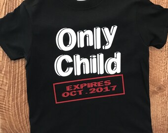 Può includere: Maglietta nera con testo bianco che recita "Only Child" in stile 3D. Sotto il testo c'è un rettangolo rosso con testo bianco che recita "EXPIRES OCT. 2017".