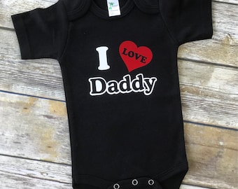 Può includere: Body nero per neonato con testo bianco che dice "I love Daddy" con un cuore rosso.