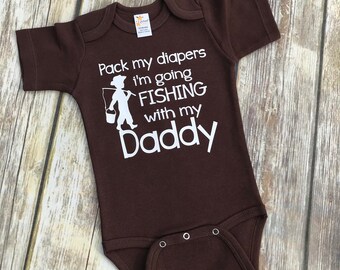 Può includere: Body da bambino marrone con testo bianco che recita "Pack my diapers I'm going fishing with my Daddy". Accanto al testo c'è una silhouette bianca di un bambino che tiene una canna da pesca e un secchio.