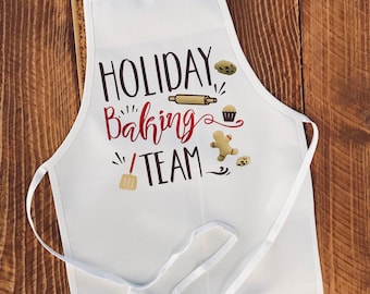 Può includere: Grembiule bianco con testo rosso e dorato che dice "Holiday Baking Team". Il testo è circondato da immagini di utensili da forno e dolcetti, tra cui un mattarello, un cupcake, un omino di pan di zenzero e una spatola.