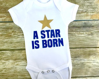 Può includere: Un body bianco per neonato con una stella dorata e il testo "A STAR IS BORN" in blu.