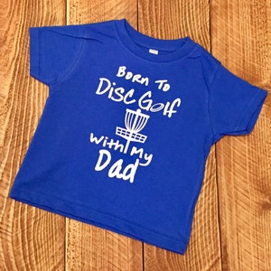 May include: A royal blue baby t-shirt with a white graphic that says "Born to Disc Golf with my Dad" and a disc golf basket.