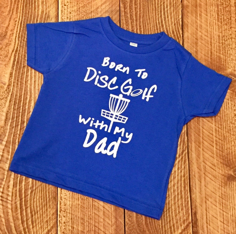 May include: A royal blue baby t-shirt with a white graphic that says "Born to Disc Golf with my Dad" and a disc golf basket.