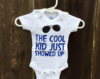 Può includere: Body da bambino bianco con grafica di occhiali da sole neri e la scritta blu "THE COOL KID JUST SHOWED UP". Il body è appeso a un gancio bianco su uno sfondo di legno.