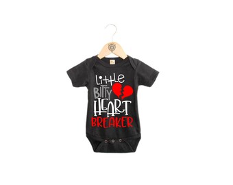 Può includere: Un body per neonato nero con una grafica bianca che dice "little bitty heart breaker" con un grafico di cuore spezzato rosso.