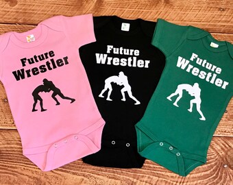 Può includere: Tre body per neonati in rosa, nero e verde con il testo "Future Wrestler" e una silhouette di due lottatori su ciascuno.