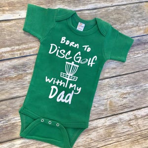 May include: Green baby onesie with white text that reads "Born to Disc Golf With My Dad" and a graphic of a disc golf basket.