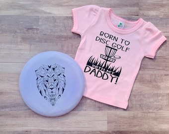 Può includere: Una maglietta rosa per neonati con la scritta "Born to Disc Golf with my Daddy!" e un'illustrazione in bianco e nero di un cestino da disc golf. Nell'immagine è presente anche un disco viola con un leone geometrico in bianco e nero.
