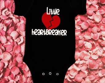 Può includere: Body nero per neonato con testo bianco che recita "Little heartbreaker" e un grafico di cuore spezzato rosso.