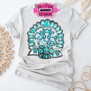 May include: White t-shirt featuring a turquoise cow design with the text "Grow 'em Show 'em". The design includes turquoise-colored stones and a banner. The shirt has rolled-up sleeves.