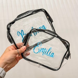 May include: Two clear plastic zippered pouches with black trim. The larger pouch features the name Barbara in blue script, while the smaller pouch features the name Emilia in blue script. Both bags are transparent and rectangular in shape.