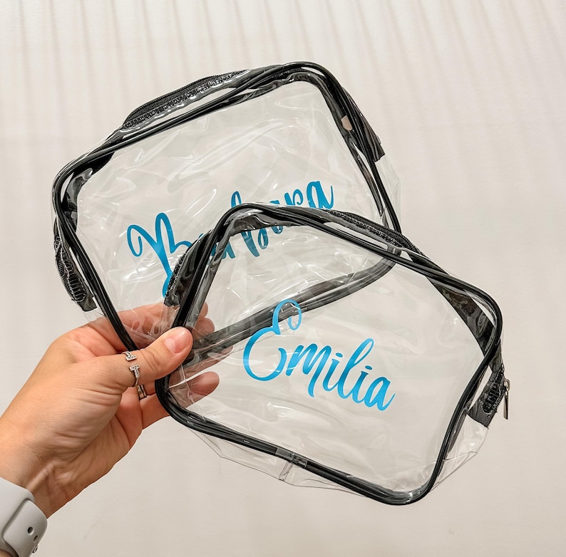 May include: Two clear plastic zippered pouches with black trim. The larger pouch features the name Barbara in blue script, while the smaller pouch features the name Emilia in blue script. Both bags are transparent and rectangular in shape.