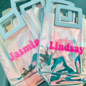 May include: Four iridescent clear plastic tote bags with blue handles. The bags are personalised with the names "Jasmin", "Lindsay", "Aria", and "Olivia" in pink letters.