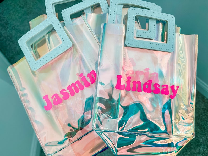May include: Four iridescent clear plastic tote bags with blue handles. The bags are personalised with the names "Jasmin", "Lindsay", "Aria", and "Olivia" in pink letters.