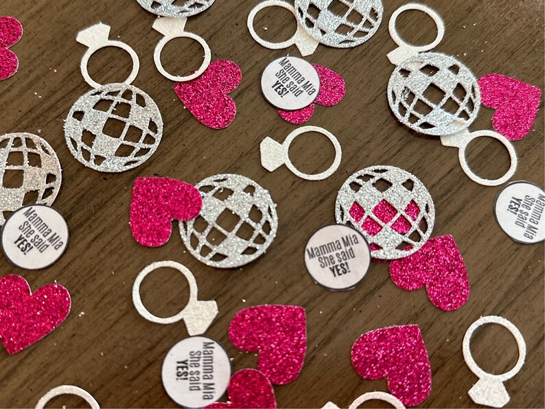 May include: Pink and silver glitter confetti with a disco ball, ring, and heart designs. The confetti also includes round circles with the text "Mamma Mia She said YES!"