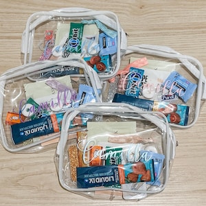 May include: Three clear plastic zippered pouches with white trim, each personalized with the name Camilla in white script lettering. The pouches are filled with various small items including Liquid I.V. packets, Orbit gum, and other assorted personal care items.