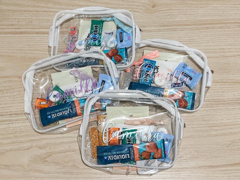 May include: Three clear plastic zippered pouches with white trim, each personalized with the name Camilla in white script lettering. The pouches are filled with various small items including Liquid I.V. packets, Orbit gum, and other assorted personal care items.