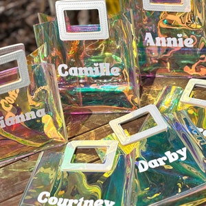 May include: Five iridescent clear plastic tote bags with white handles and white text. The bags are labelled "Camille", "Annie", "Gianna", "Darby", and "Courtney".