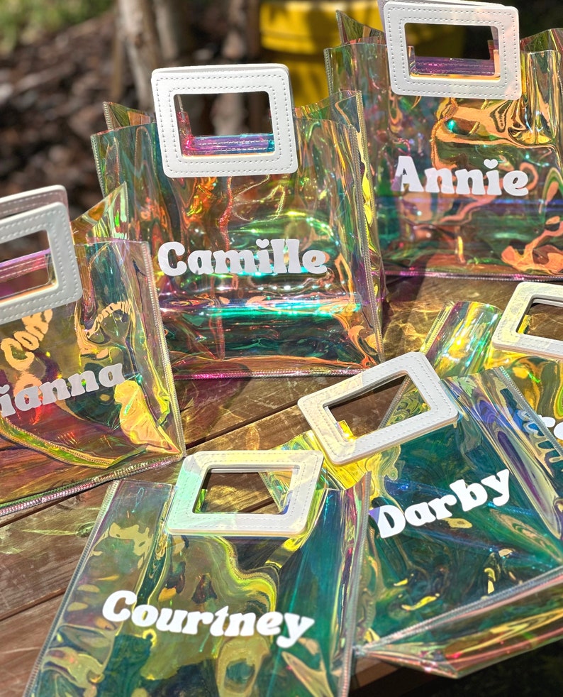 May include: Five iridescent clear plastic tote bags with white handles and white text. The bags are labelled "Camille", "Annie", "Gianna", "Darby", and "Courtney".