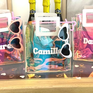 May include: Three clear plastic bags with pink handles and white frames. Each bag contains a pair of heart-shaped sunglasses and a personalised name tag. The names are "Laure", "Camille", and "Darby".