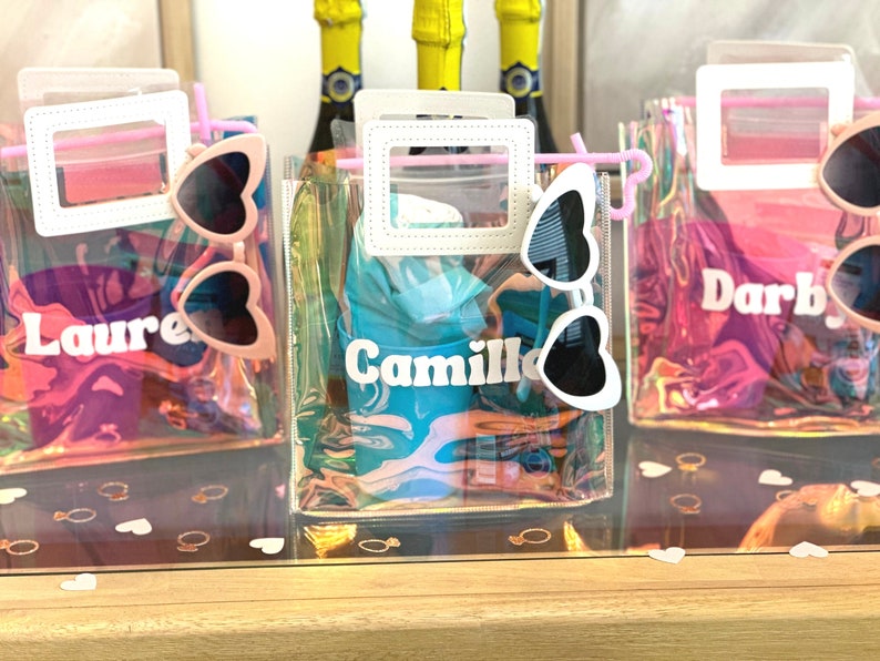 May include: Three clear plastic bags with pink handles and white frames. Each bag contains a pair of heart-shaped sunglasses and a personalised name tag. The names are "Laure", "Camille", and "Darby".