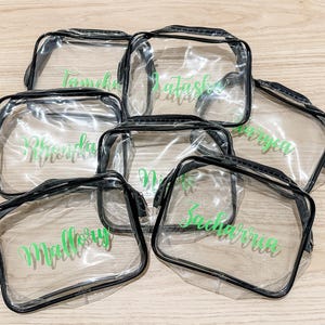 May include: A collection of six rectangular clear plastic pouches with black trim and zippers. Each pouch features a unique name printed in a vibrant green script font on the front surface, including names like Mallory, Zacharria, and Rhonda.