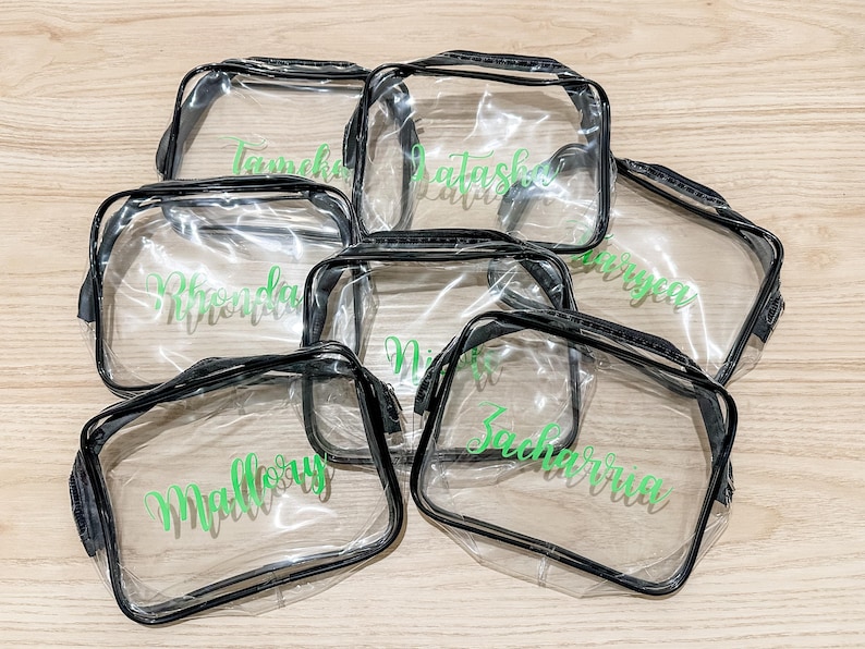 May include: A collection of six rectangular clear plastic pouches with black trim and zippers. Each pouch features a unique name printed in a vibrant green script font on the front surface, including names like Mallory, Zacharria, and Rhonda.