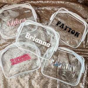 May include: Four rectangular clear plastic pouches with white zippers. Each pouch features a personalized name printed on the front: Adelina in light pink, Payton in black, Brianna in white, Reese in hot pink, and Cassidy in silver glitter.