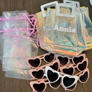 May include: A set of party favours including clear plastic bags with pink straws, clear plastic bags with the text "You're the Best Brianna" printed on them, clear plastic totes with an iridescent finish and the text "Warren Annie" printed on them, and pink heart-shaped sunglasses.
