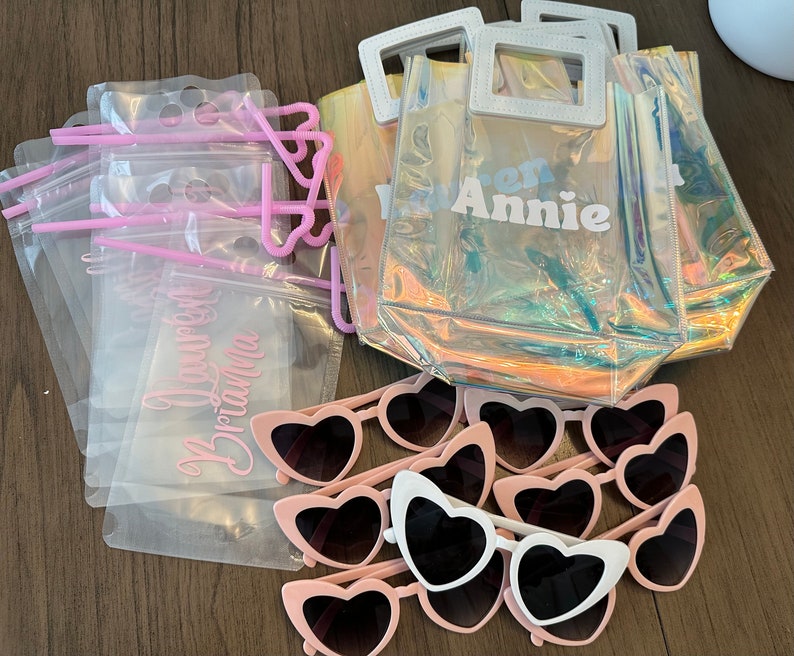 May include: A set of party favours including clear plastic bags with pink straws, clear plastic bags with the text "You're the Best Brianna" printed on them, clear plastic totes with an iridescent finish and the text "Warren Annie" printed on them, and pink heart-shaped sunglasses.