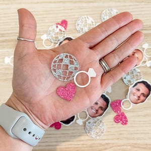 May include: A collection of silver glitter confetti featuring a disco ball, a ring, and hearts. The confetti is perfect for a wedding or engagement party.