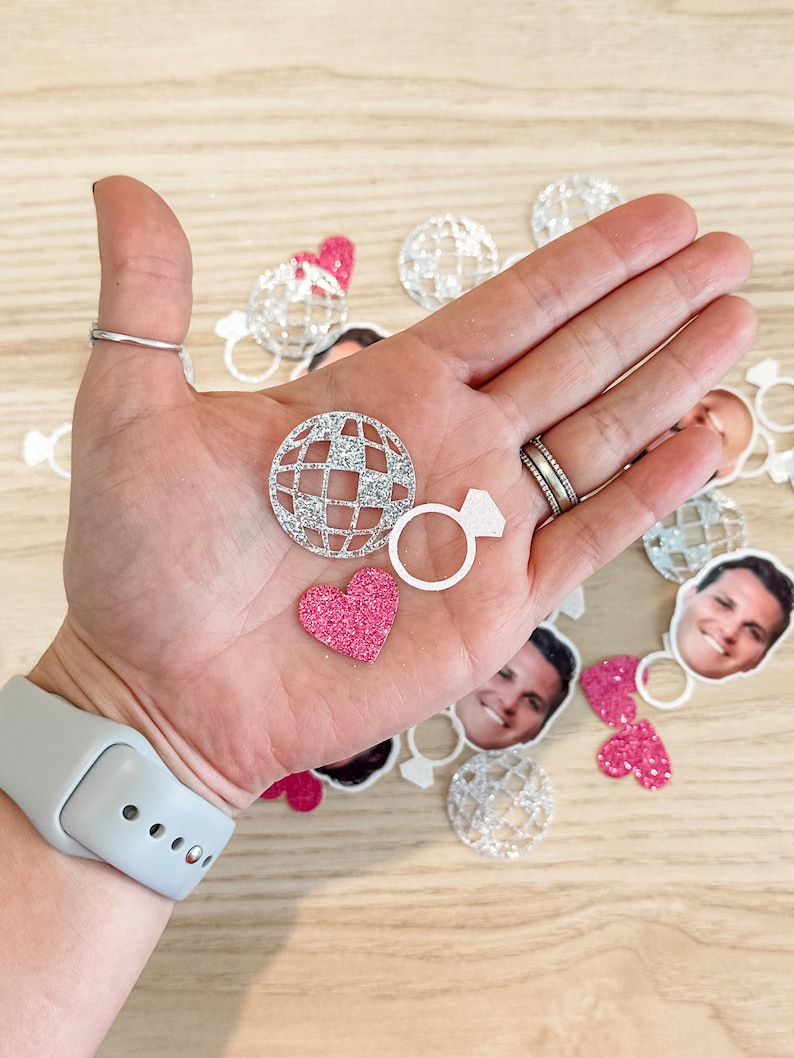 May include: A collection of silver glitter confetti featuring a disco ball, a ring, and hearts. The confetti is perfect for a wedding or engagement party.
