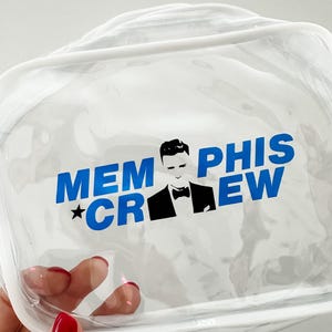 May include: A clear, rectangular vinyl pouch with white trim and a zipper closure. The front features blue text that reads MEMPHIS CREW, with a black and white graphic of a person in a tuxedo jacket and bowtie positioned between the words.